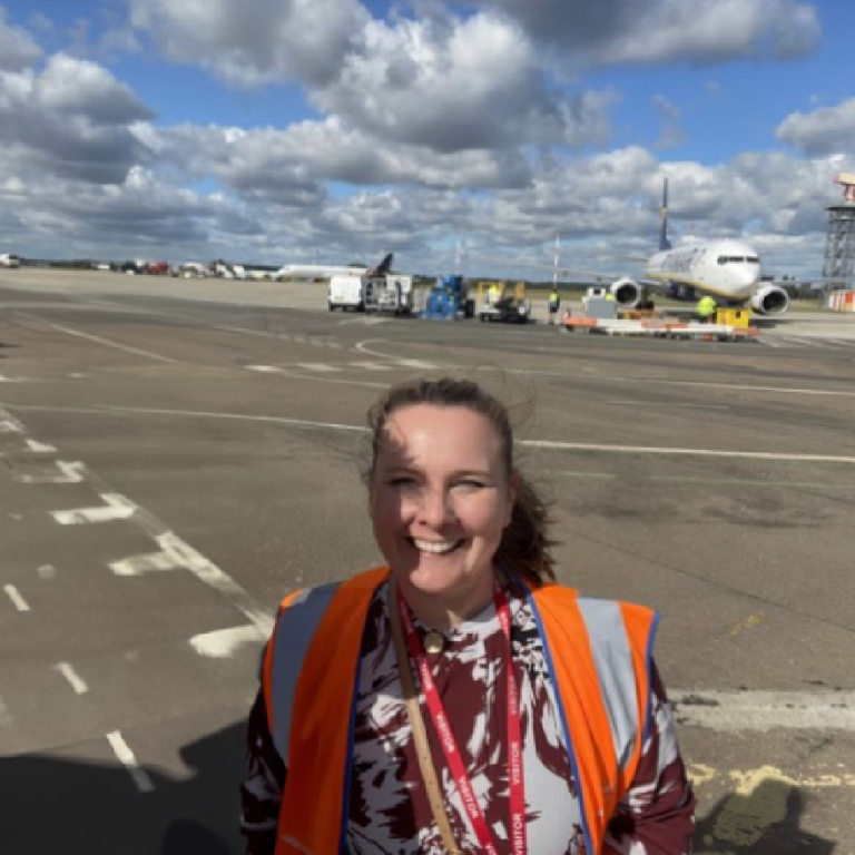 Headshot of Clair Bond standing on a runway at a comercial airport wearing an orange high visibility vest over casual clothing, there are planes being loaded in the background